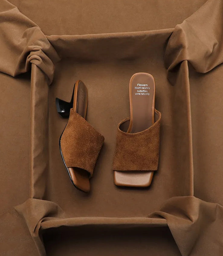 A pair of women's heeled slide sandals in a warm brown suede-like material, displayed inside a brown fabric-lined box. One sandal is shown from the side, revealing a black heel, and the other is shown from the top, revealing a square toe and a light beige insole with the brand name 'Flowers Direct'.