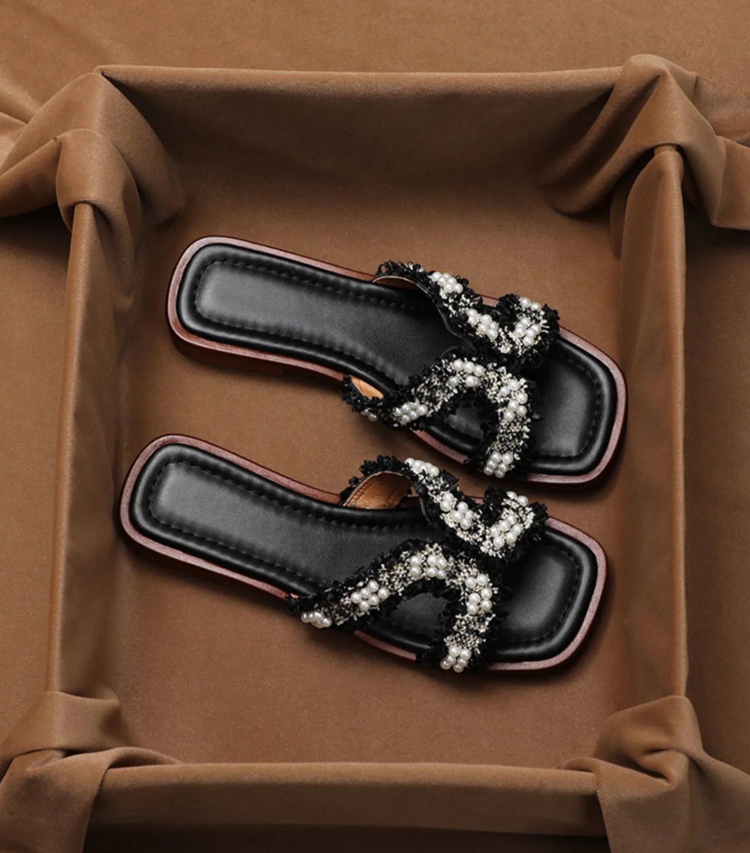 A pair of black leather slide sandals with wide, ornate straps heavily embellished with white pearls and silver rhinestones, resting inside a brown fabric-lined box.