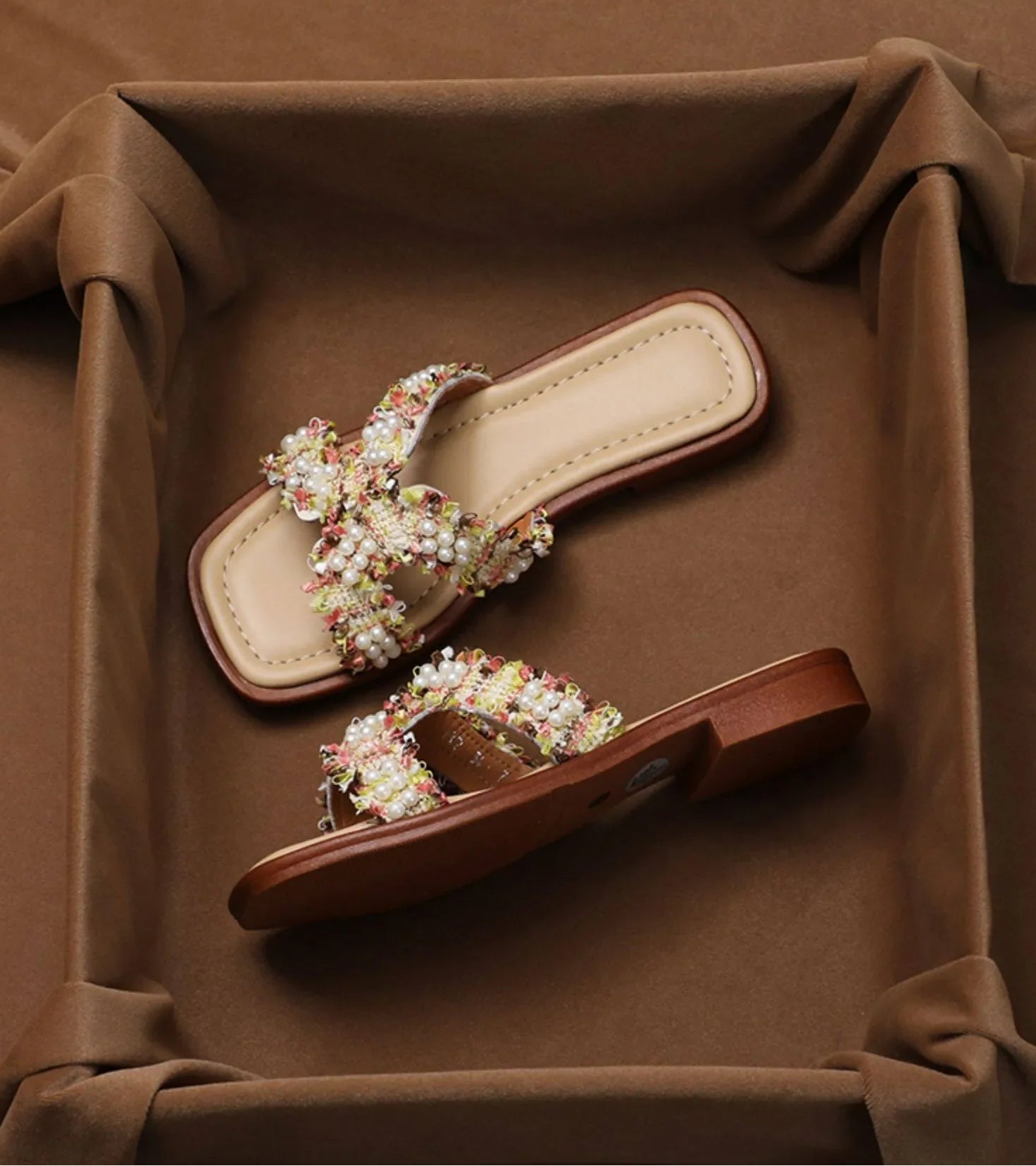 A pair of women's flat slide sandals with wide, embellished straps, displayed inside a brown fabric-lined box.