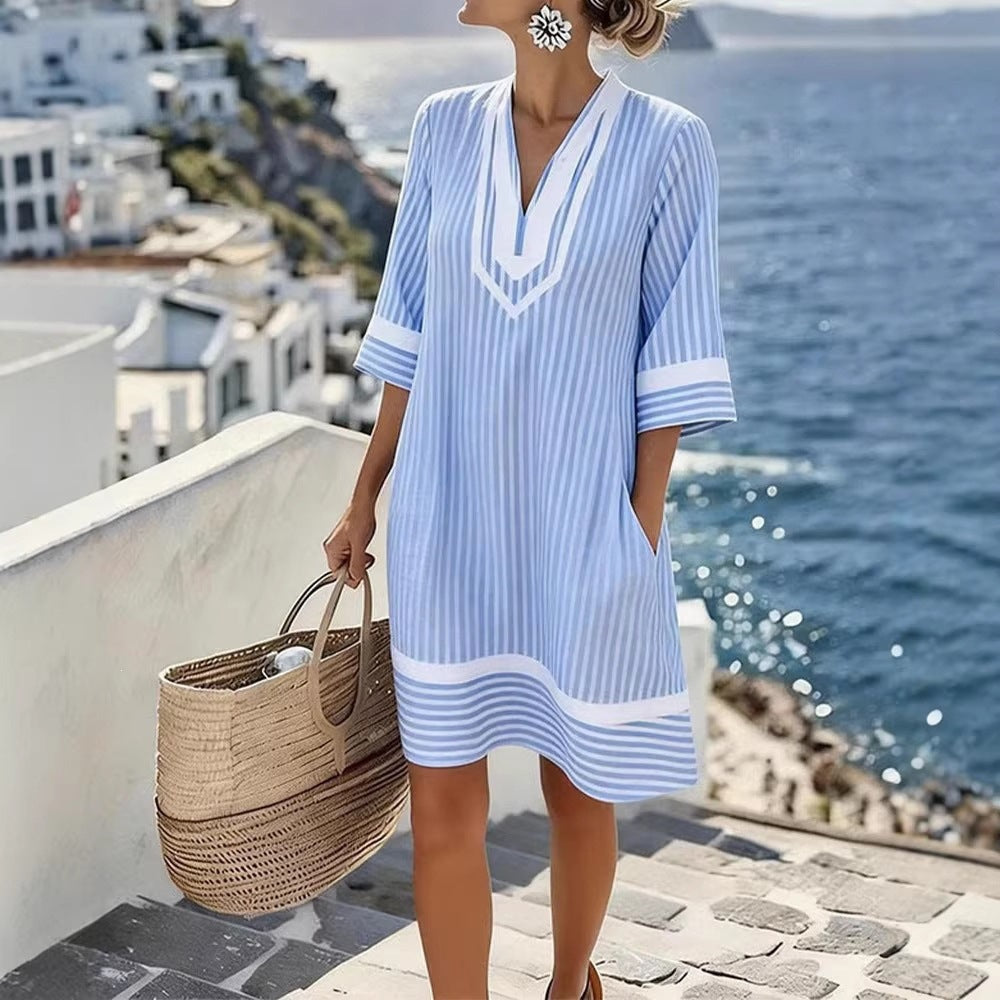 A woman wearing a light blue and white striped beach cover-up dress with a V-neck and elbow-length sleeves, standing outdoors by the sea and holding a woven straw tote bag.