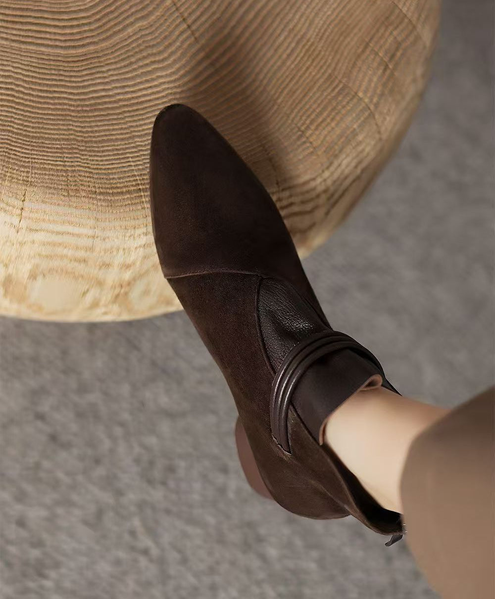 A dark brown ankle boot with a pointed toe and a decorative strap detail on the side, shown worn on a foot resting on a woven straw stool.