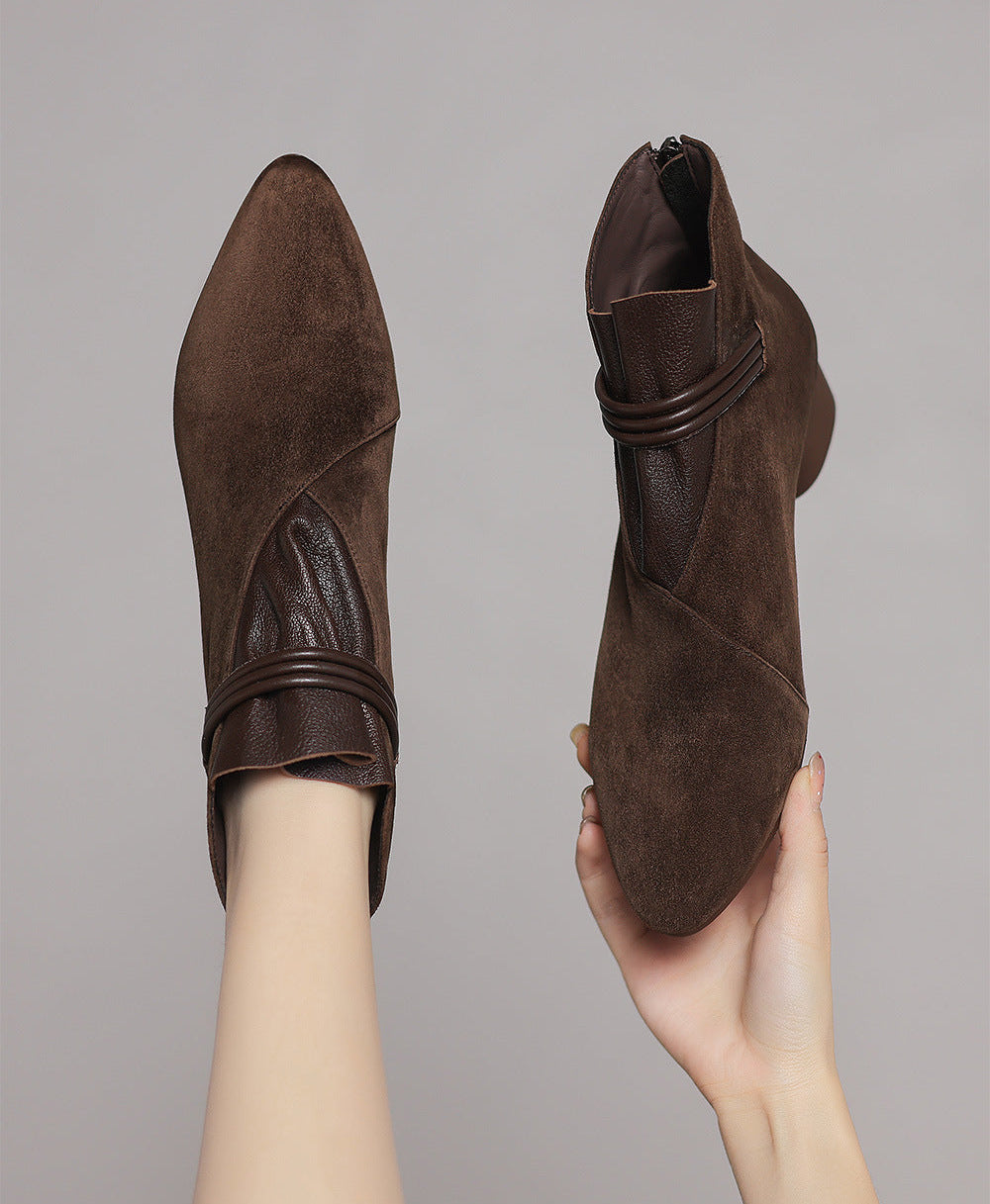 A pair of dark brown suede ankle boots with faux leather strap details, shown with one boot worn on a foot and the other held in a hand against a solid gray background.